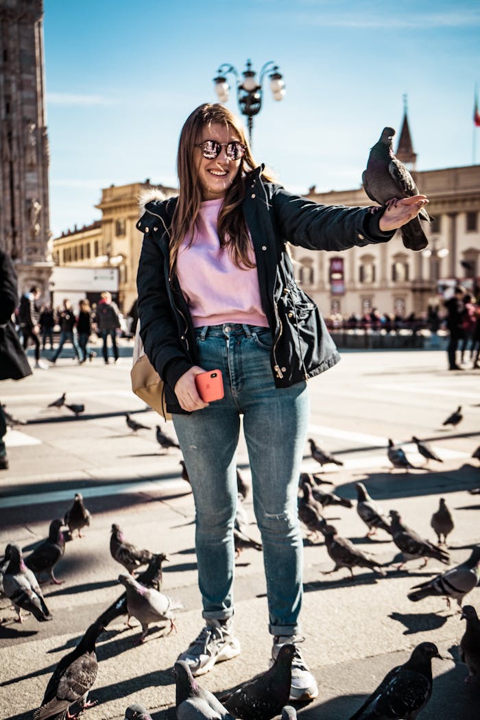 A young woman enjoys a sunny day, feeding pigeons in a bustling city square, capturing a lively urban moment.